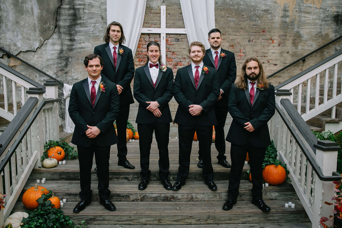 Six men in black suits with burgundy ties stand on wooden steps decorated with pumpkins and greenery. A white draped cross and exposed brick wall are in the background, giving a rustic, autumn wedding vibe.