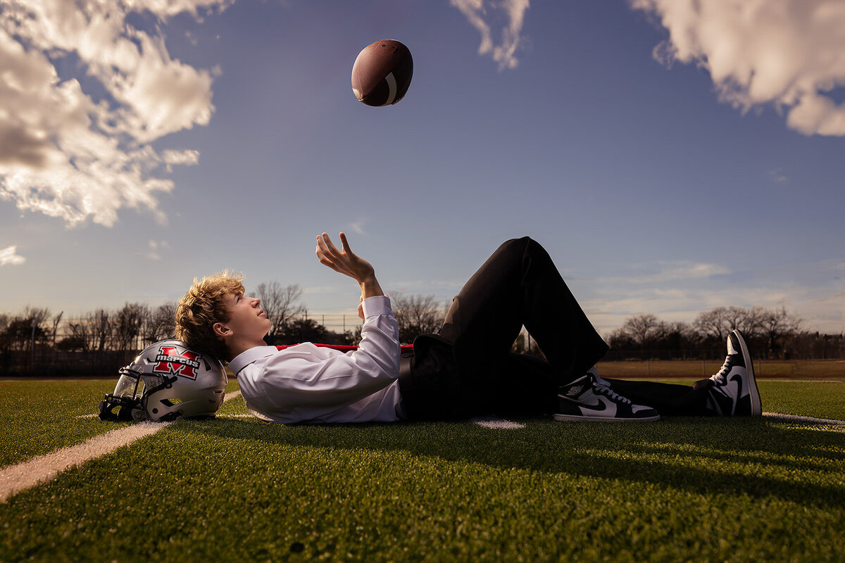 Senior in a suit, lounging on the football field with a helmet by his side and a football in the air. Capturing the mix of athletic and professional spirit for a unique senior photoshoot in Dallas.
