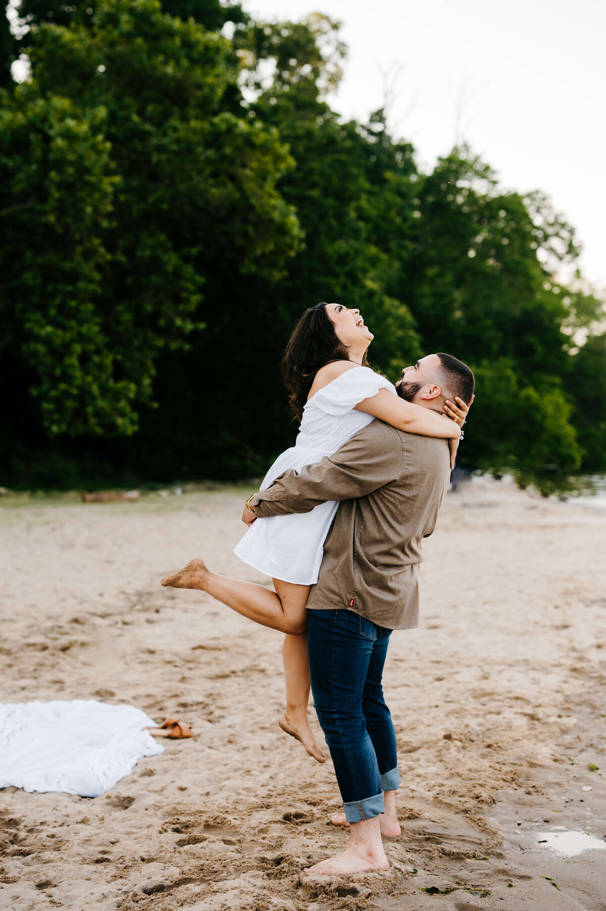 beach engagement photos at woodbridge lake with man picking up woman in a white dress as she throws her head back and laughs