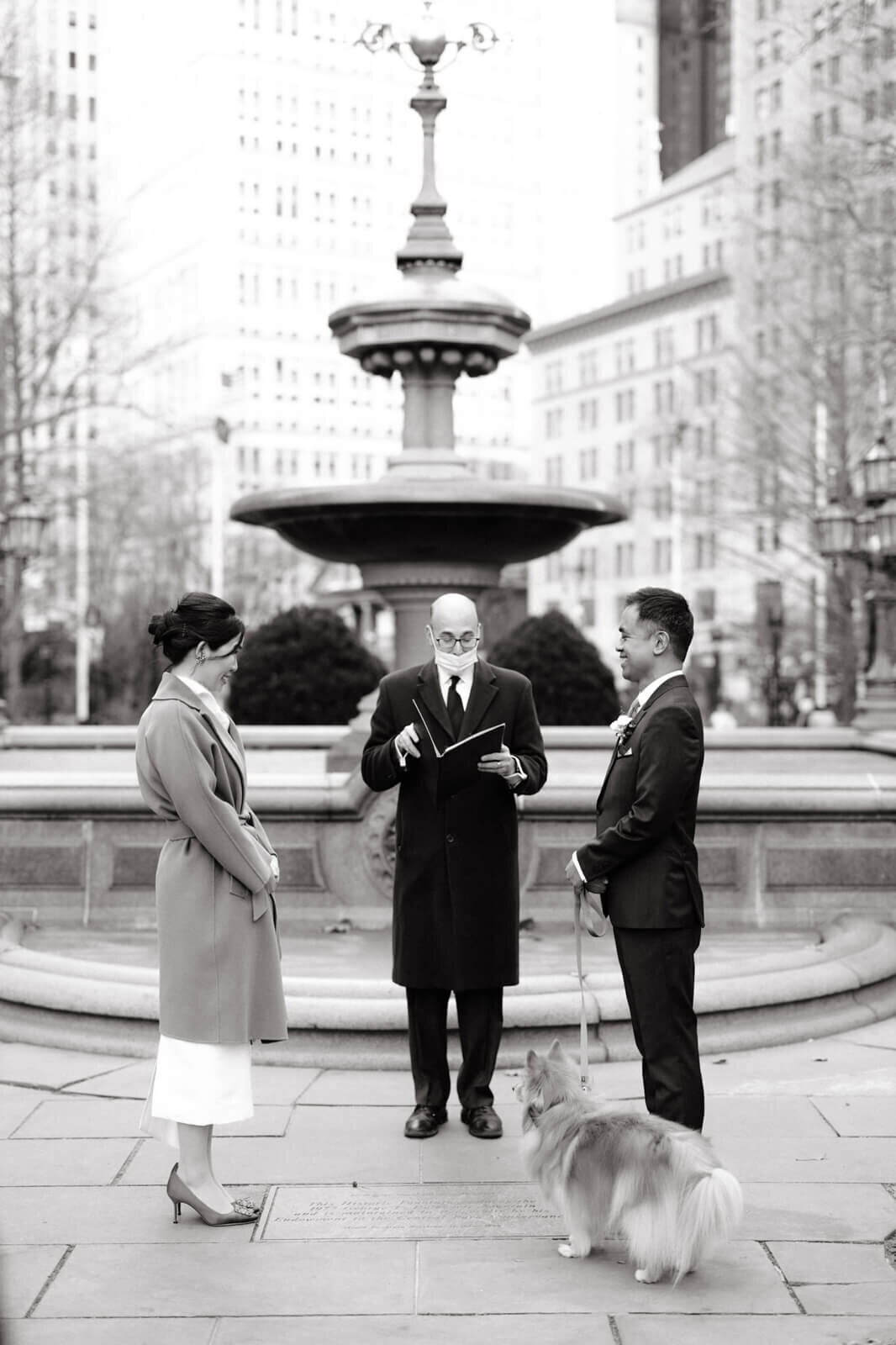 Black and white photo of the bride and groom, with their dog, saying their vows to a wedding officiator in front of a fountain.