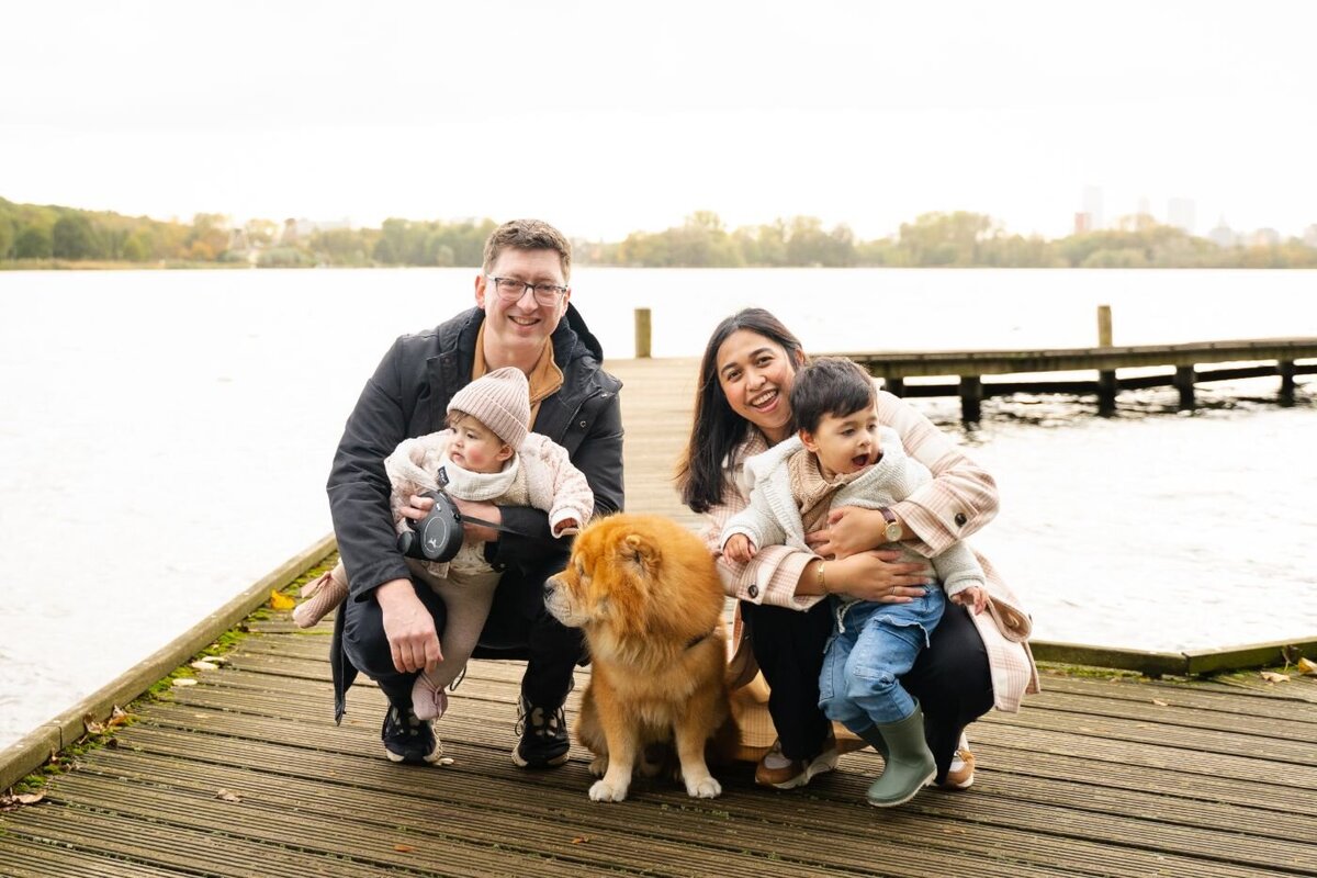 Family with dog by lake – Smiling parents with two small children and a fluffy brown dog posing on a lakeside wooden deck with autumn colors in the background.