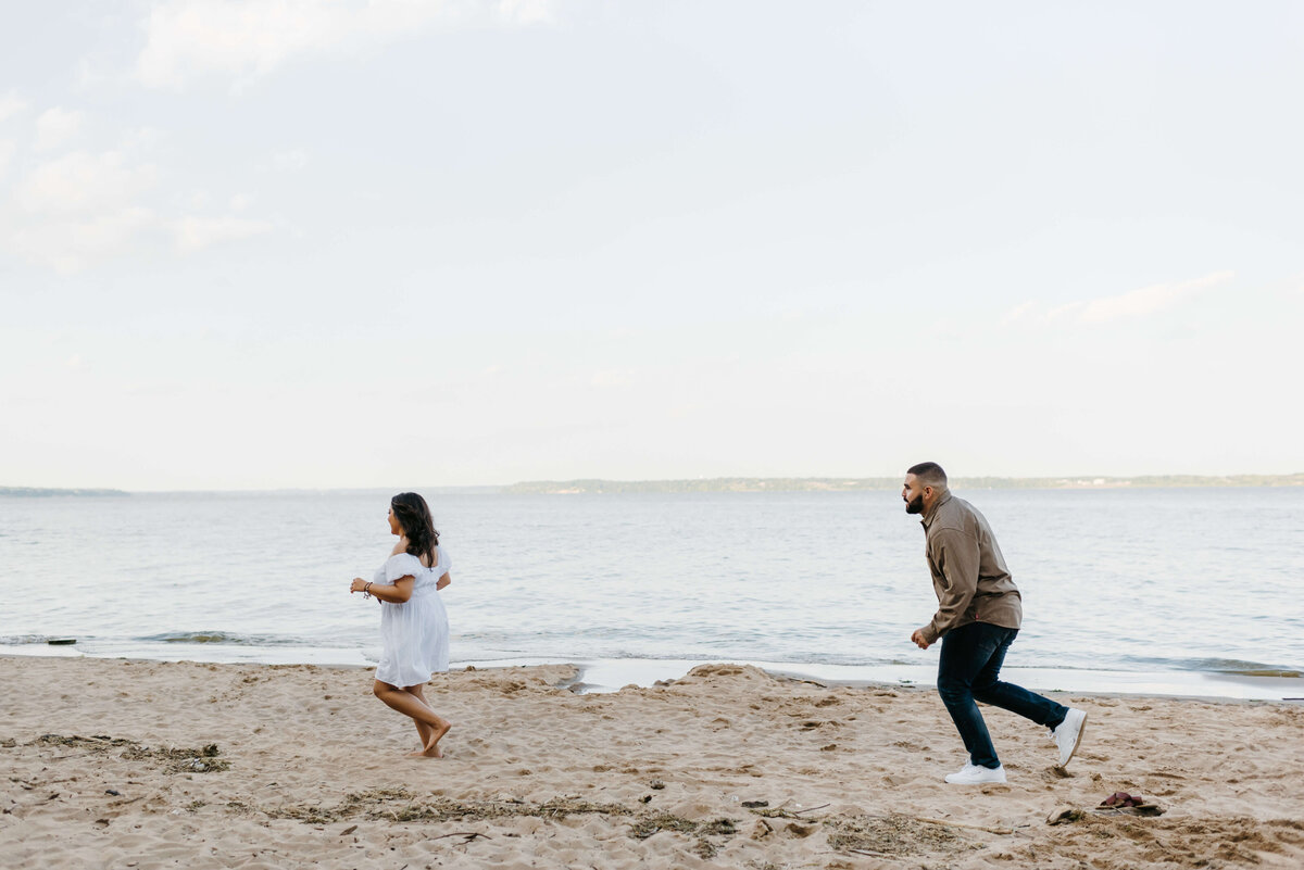 candid engagement phot of man chasing his fiance on the beach as they play together on the shore of woodbridge lake