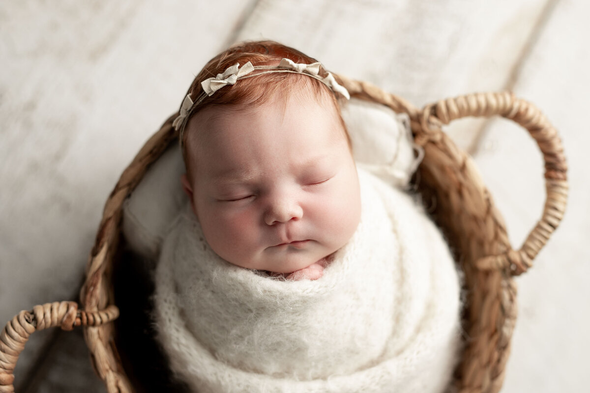 Studio newborn photography - sleeping newborn baby girl with red hair wrapped in cream with a delicate petite headband with bows. Baby is wrapped in a cream swaddle.