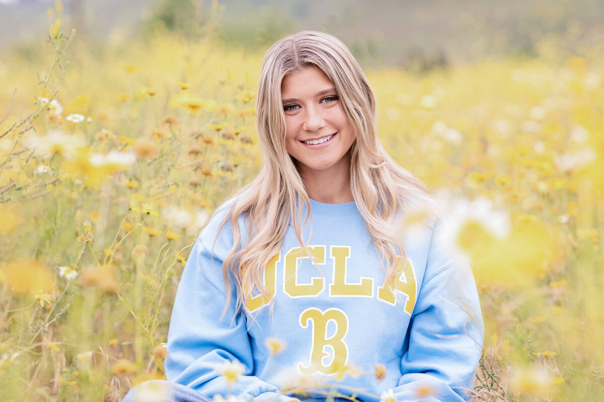 A person with long hair, wearing a blue UCLA sweatshirt, sits in a field of yellow flowers, smiling at the camera. The background is a blurred, natural setting.