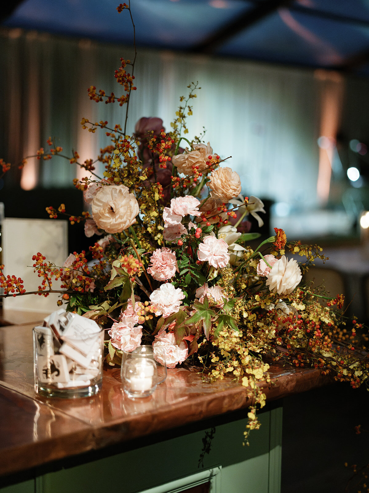 Trailing bar meadows with florals in hues of purple, ruby, copper, gold, and rust for fall wedding reception. Sculptural product clustered floral design for fall wedding. Product clustered floral statements in a tasteful fall color palette of copper, ruby, gold, burgundy, rust, and lavender. Orchids, bittersweet berries, butterfly ranunculus, and tulips are highlighted. Unique floral design with flower forward large scale installations. Design by Rosemary & Finch Floral Design in Nashville, TN.