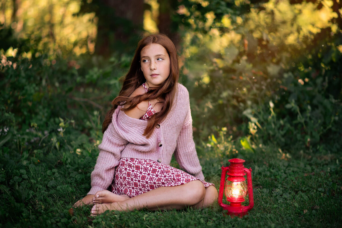 A little girl with red hair sits in the grass with a glowing lantern by her side looking off into the distance. Captured in New Brunswick by Brittany Danielle