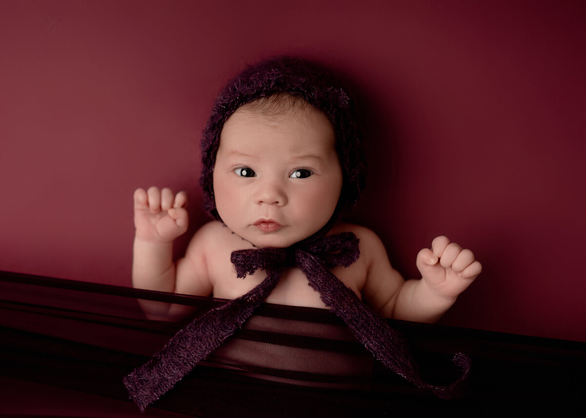 A newborn baby in a purple bonnet with large bow lays on a bed with eyes open and arms up