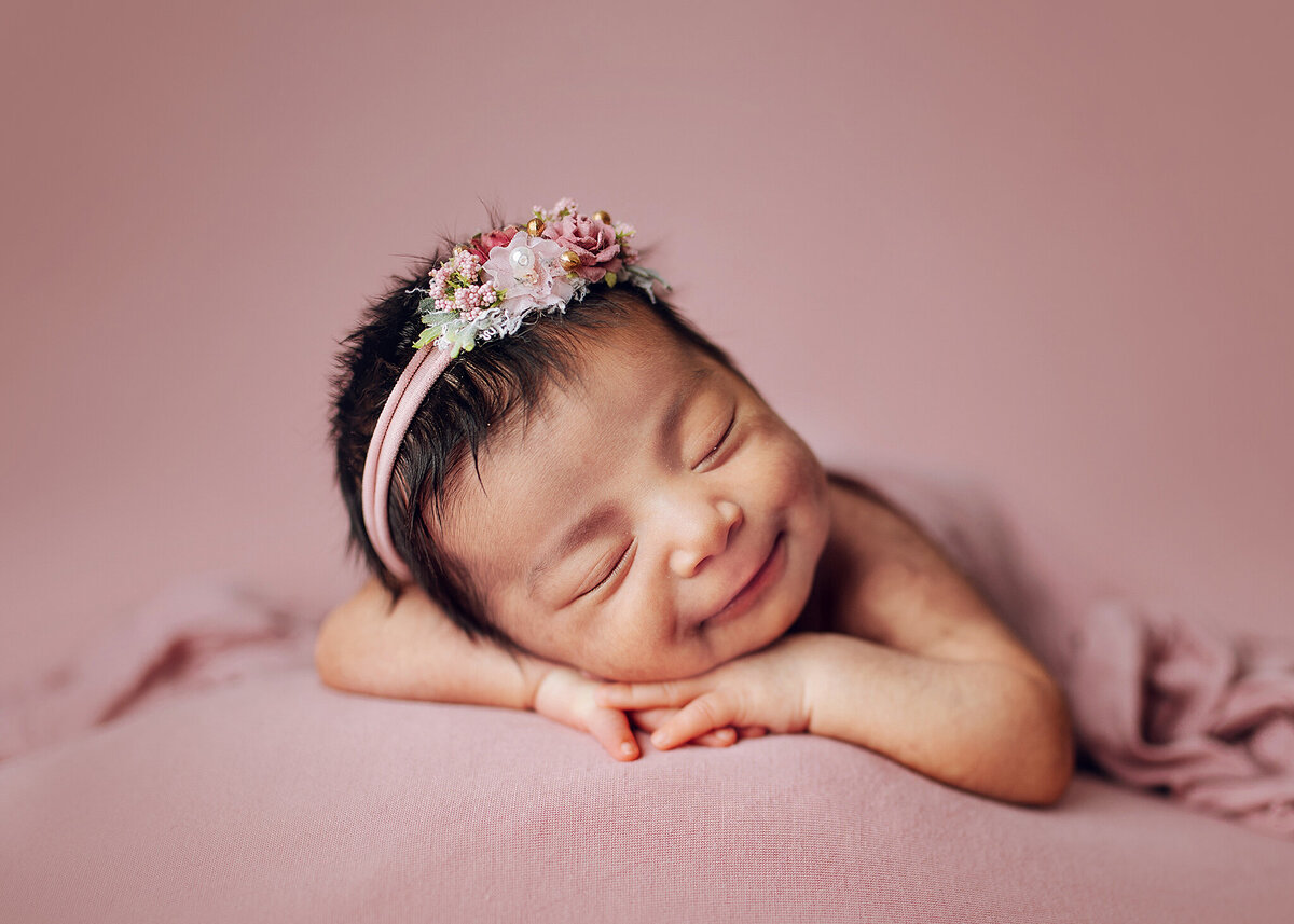 newborn girl posed on her hands wearing a pink headband laying on a pink fabric and covered by a pink wrap