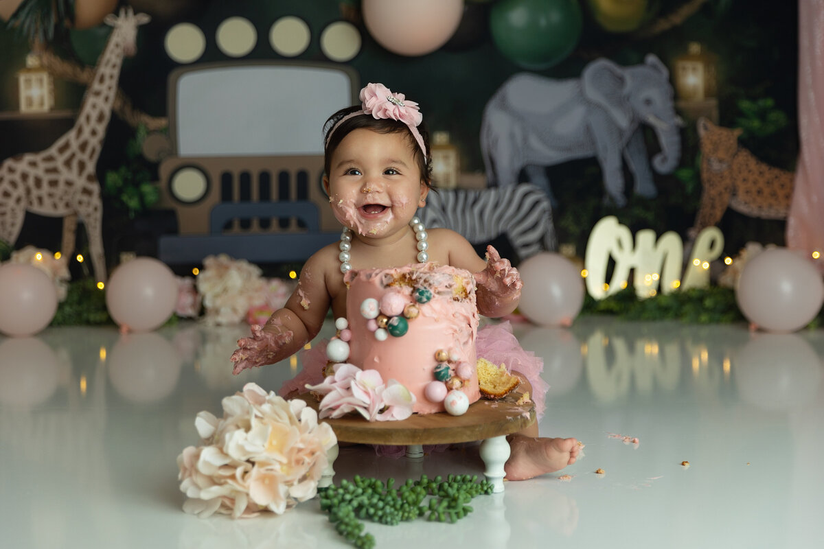 A happy toddler girl wearing a pearl necklace smiles big while eating a cake by herself in a studio