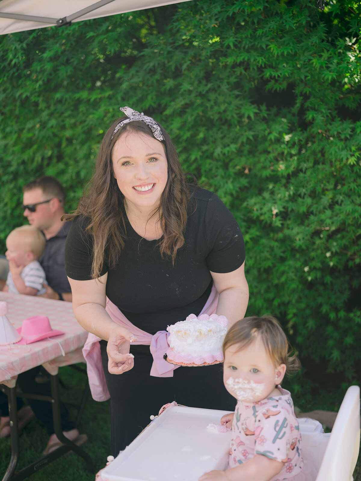 Mom smiling at the camera while helping her baby girl with the smash cake at a first birthday party in the San Francisco Bay Area.