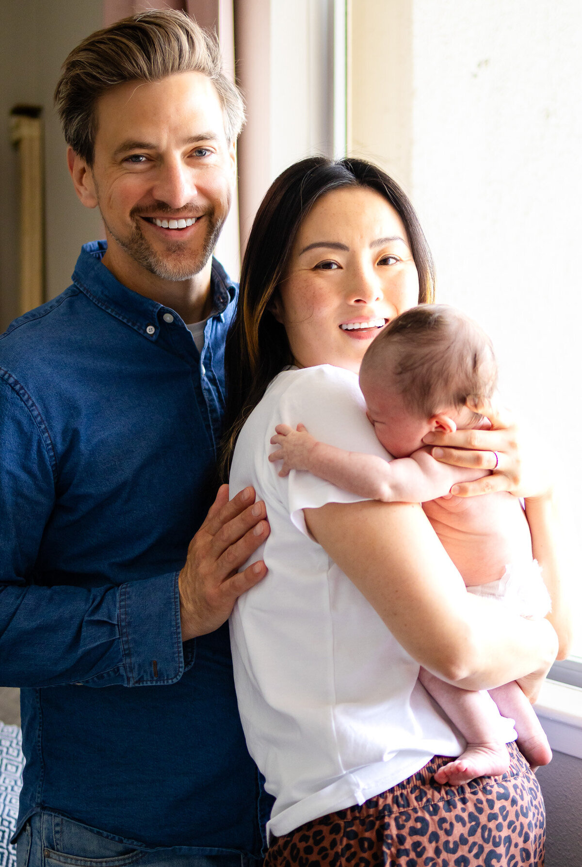 Family portrait at the window with father smiling and mother holding baby on her shoulder – natural light photo by San Francisco newborn photographer.