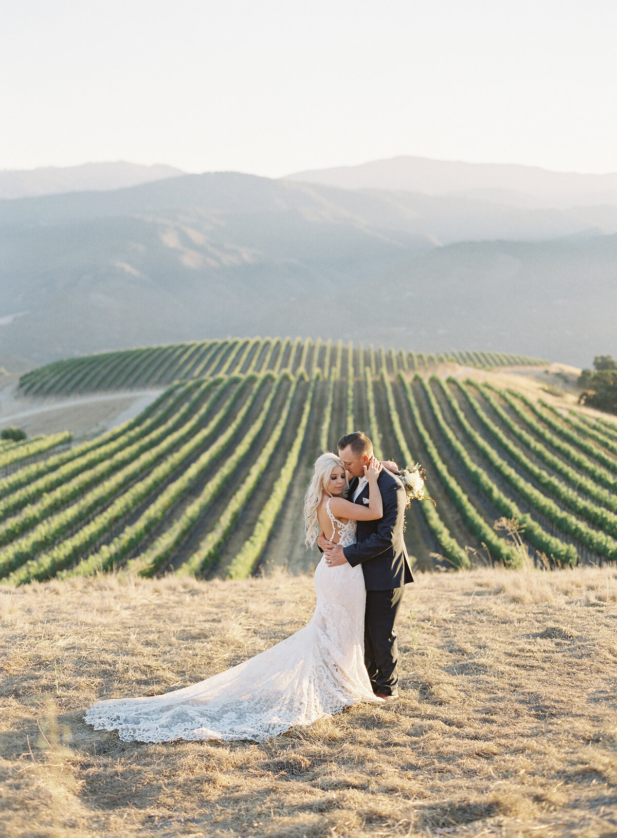 A couple sharing a romantic kiss on top of a mountain at Holman Ranch, overlooking the rolling vineyards of carmel and Napa Valley. The breathtaking view, paired with the golden light of sunset, creates an unforgettable and timeless wedding moment.