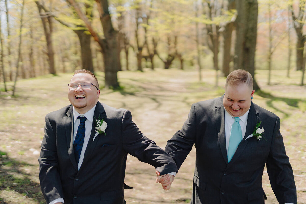 Gay couple holding hands walking through forest at Rochester NY wedding