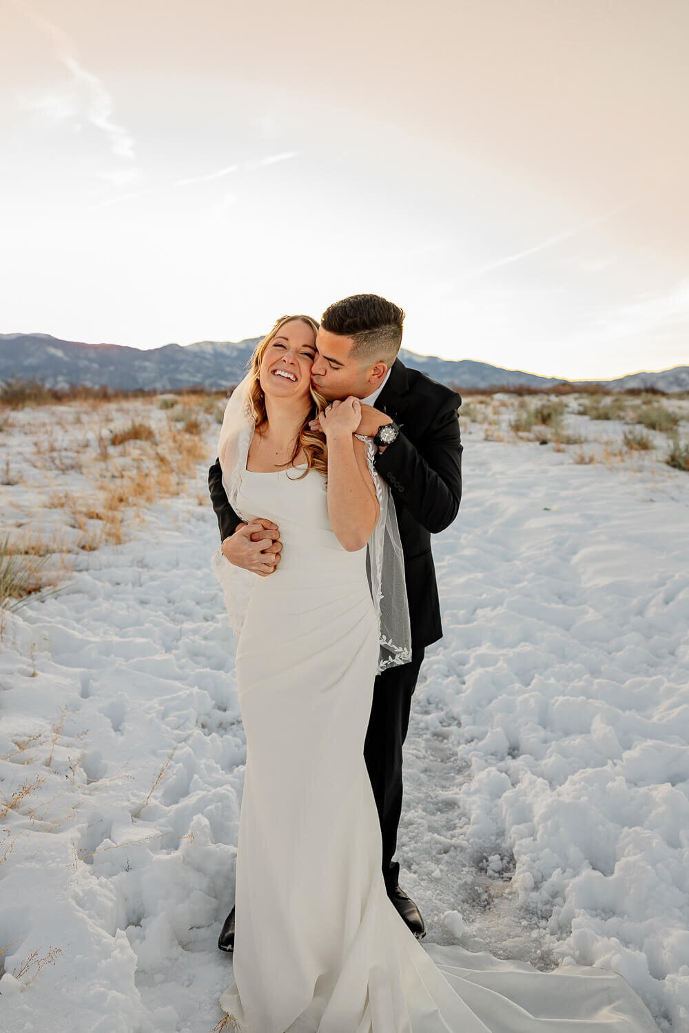 Groom hugging bride from behind and kissing her in the snow. Captured by Michelle Betz Photography