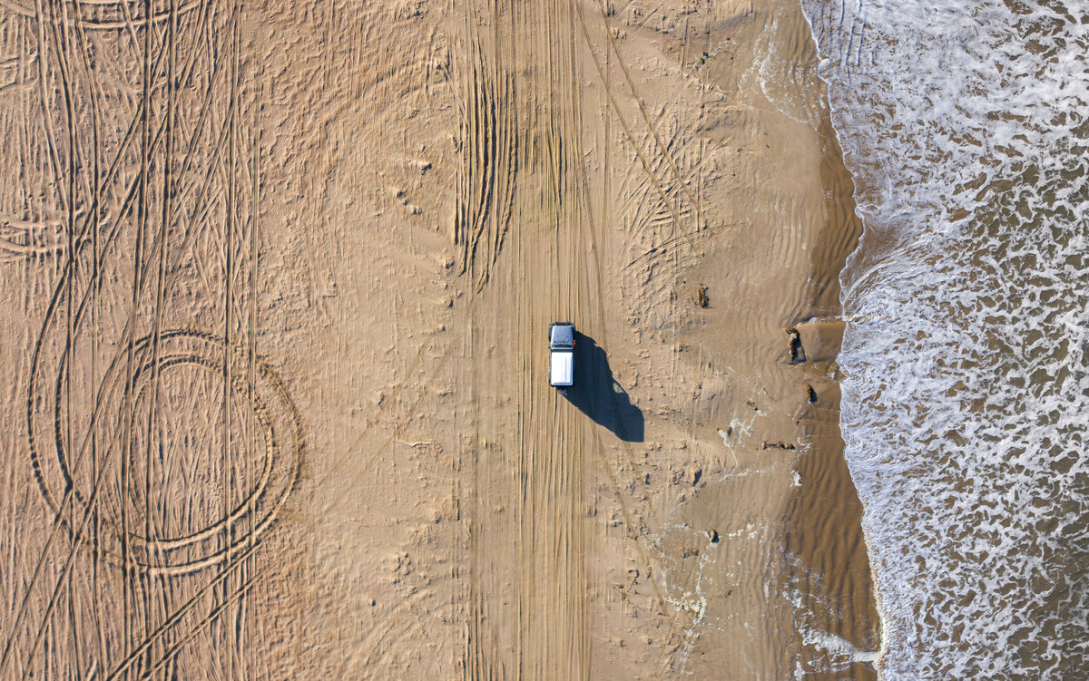 Aerial view of a Jeep driving along the sandy shores of Pismo Beach, California, leaving tire tracks in the sand as part of a luxury overland adventure along the Pacific Coast