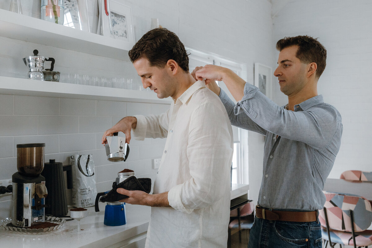 A man fixing his partner's collar as the other makes coffee.