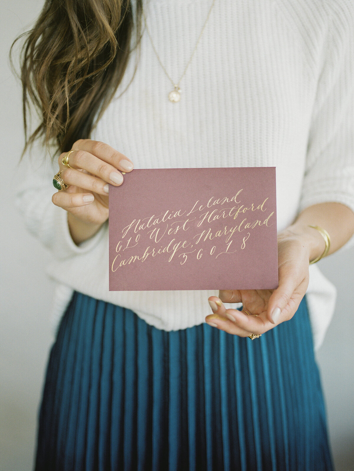 woman with cream sweater, blue pleated skirt holds purple stationery envelope with gold calligraphy ink