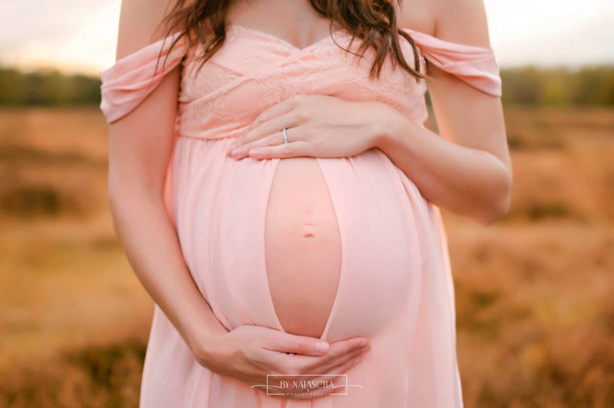Zwangere vrouw vanaf de voorzijde een close-up van haar zwnagere buik die ze vasthoudt met een hand boven en onder haar buik en de jurk een beetje open zodat de navel zichtbaar is