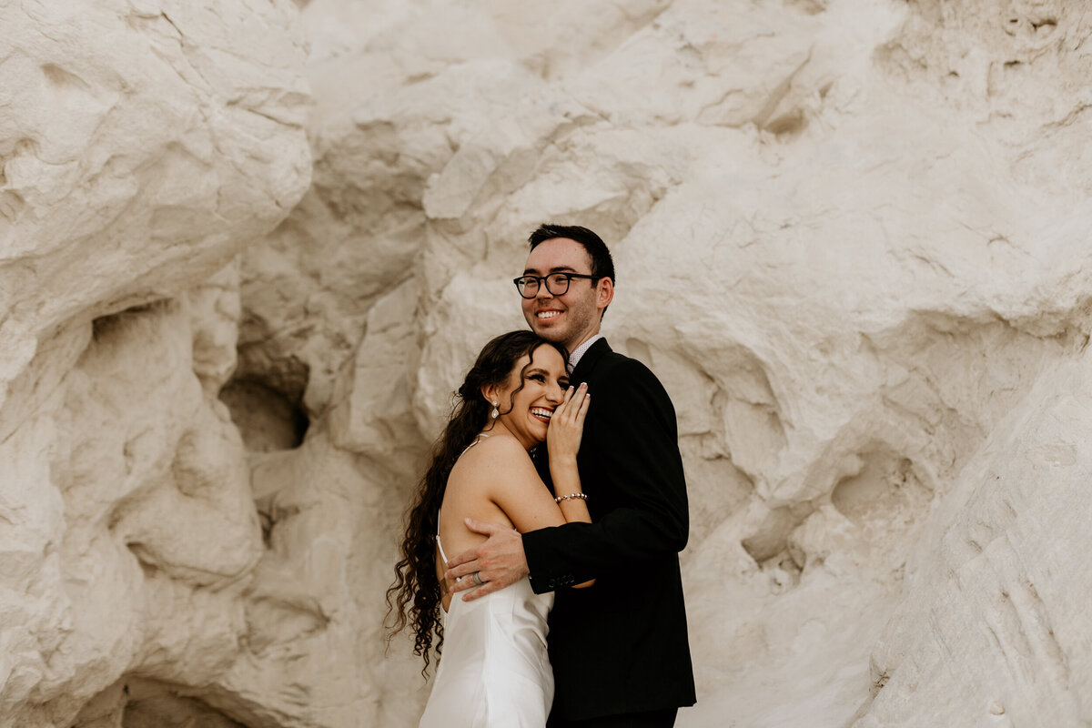 bride and groom hugging and looking off in front of a white rock