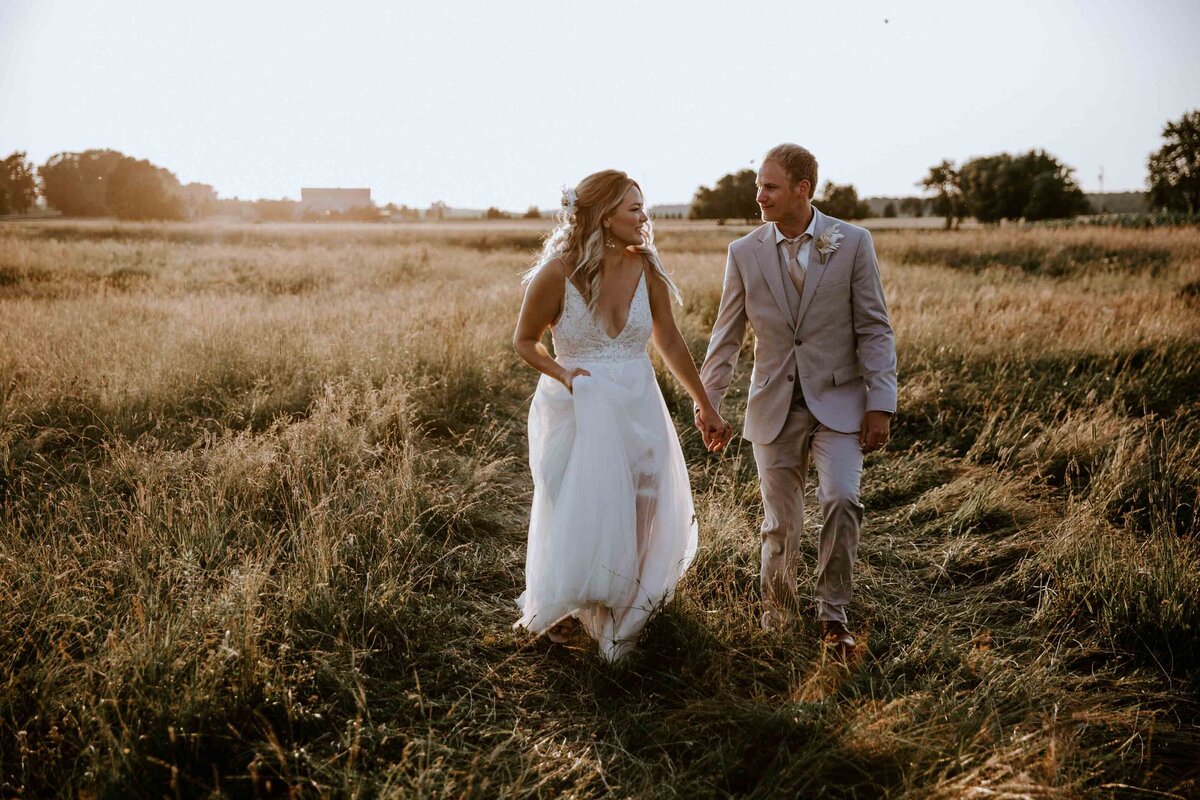 Bride and Groom walking through Exeter, Ontario field on their wedding day for photos are golden hour. They are looking at each other and holding hands. Soft golden light and sun haze surround them.