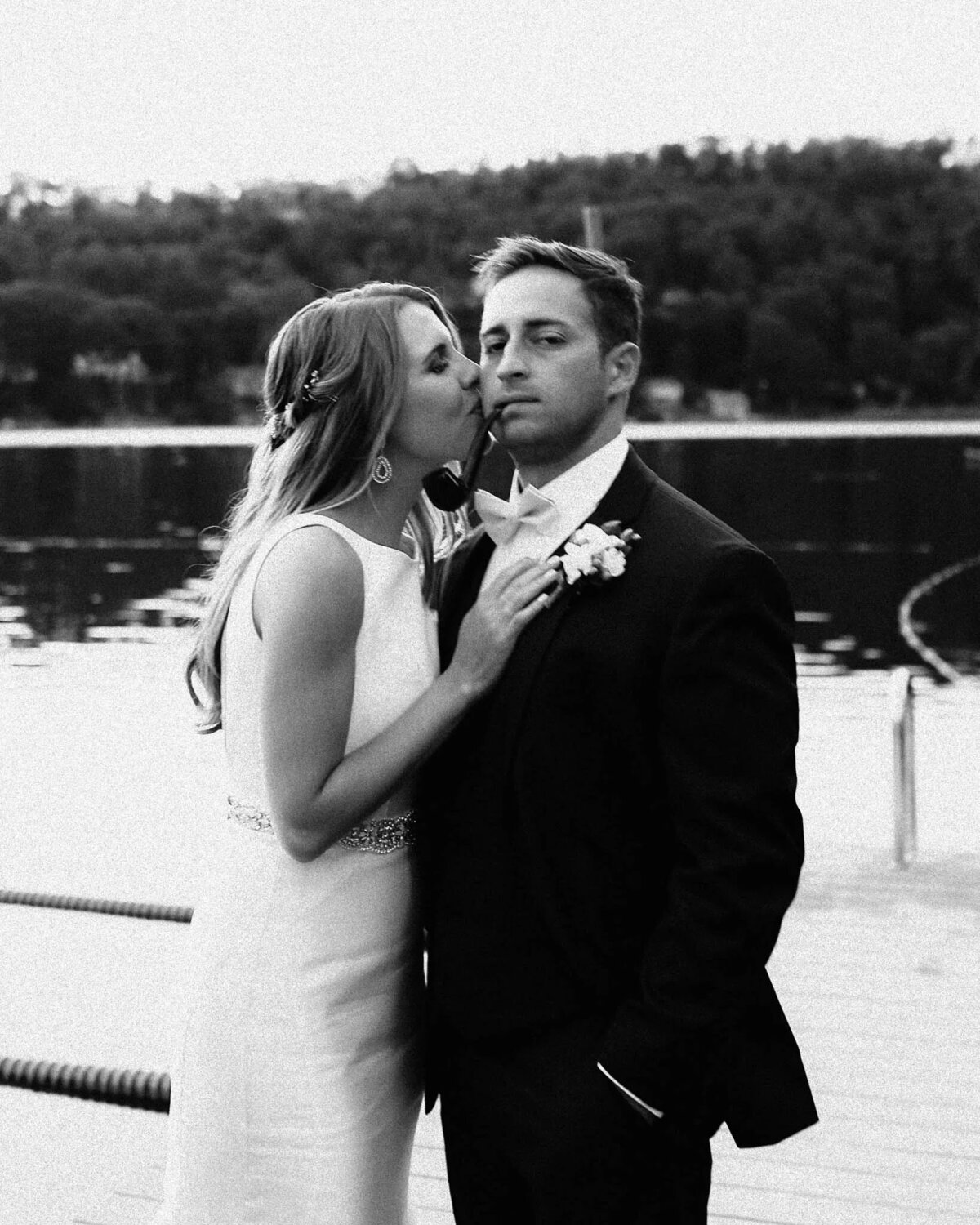 A bride in a white dress kisses a serious-looking groom on the cheek by a calm lake, with trees in the background—captured beautifully by a film photographer NJ couples trust for timeless moments.