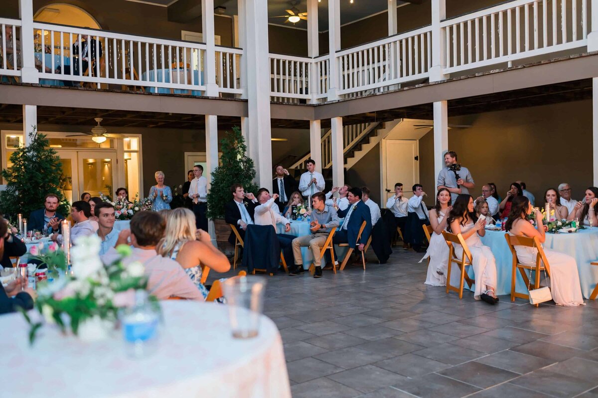 A group of wedding guests in formal attire sit at round tables outdoors under a large, two-story veranda, attentively watching a speaker during a reception.