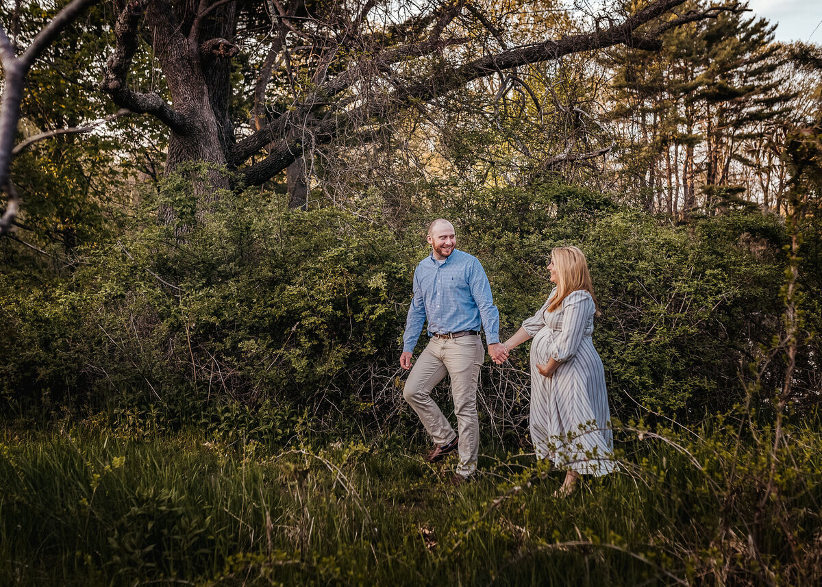 husband leading pregnant wife through woods at maternity photography session at carey cottage in portsmouth nh
