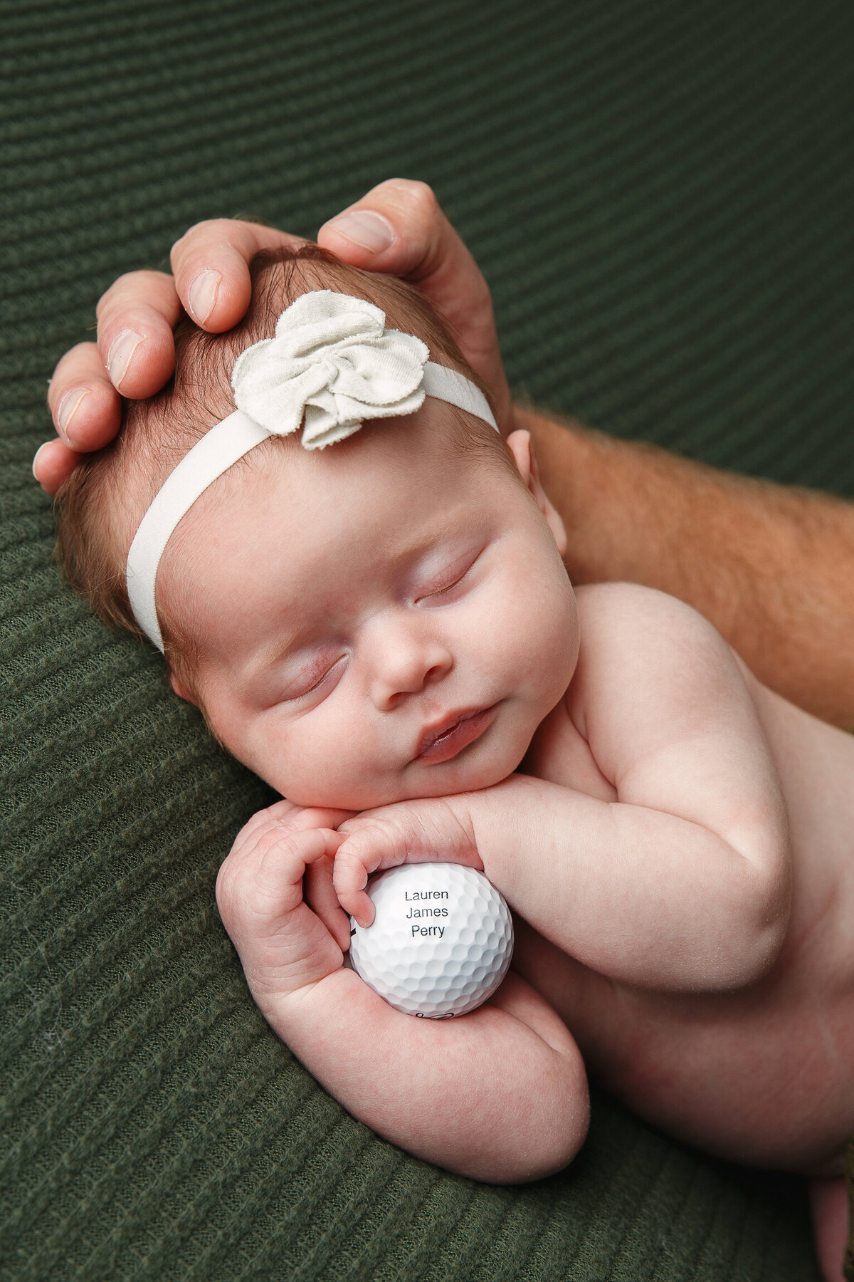 Portrait of an infant girl holding a golf ball and showing dad's hand on her head