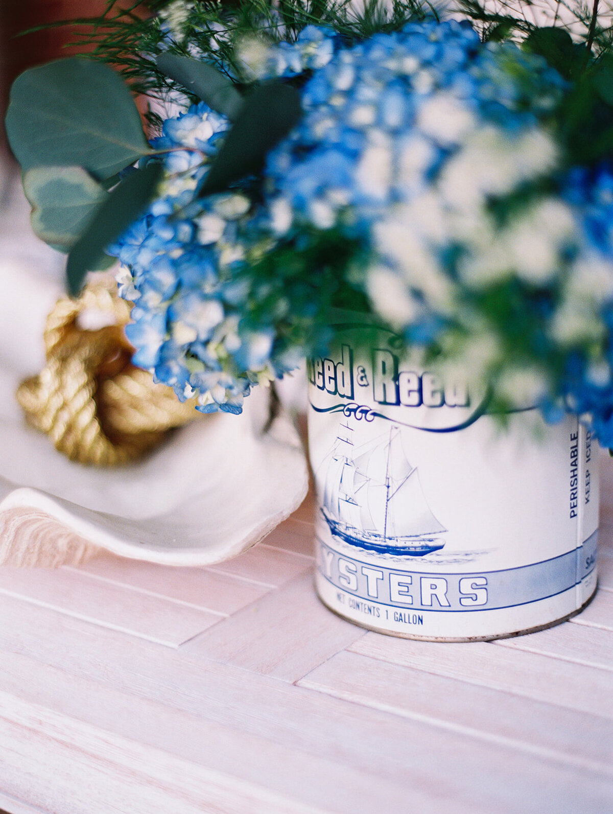 The picturesque setting for this elegant scene is The Hamptons, New York, at a waterfront estate tented wedding captured by photographer Bonnie Sen. The decor showcases a creative touch with a can of Rappahannock oysters repurposed as a vase, brimming with a lively bouquet of blue and white flowers accented with lush greenery. Adorning the can is a charming illustration of a sailing ship, adding to the nautical theme. Completing the arrangement are a dainty nautical knot and a delicate white seashell placed nearby on the pristine white surface. Additionally, East West Films has been added as videographers to the event.