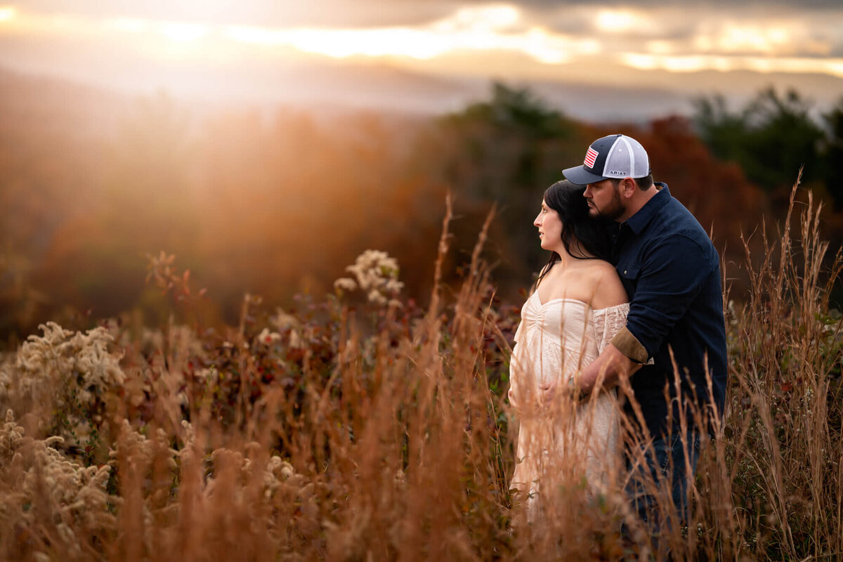 An expecting couple stand in the tall grass and cradle their baby bump  as the sun sets behind them