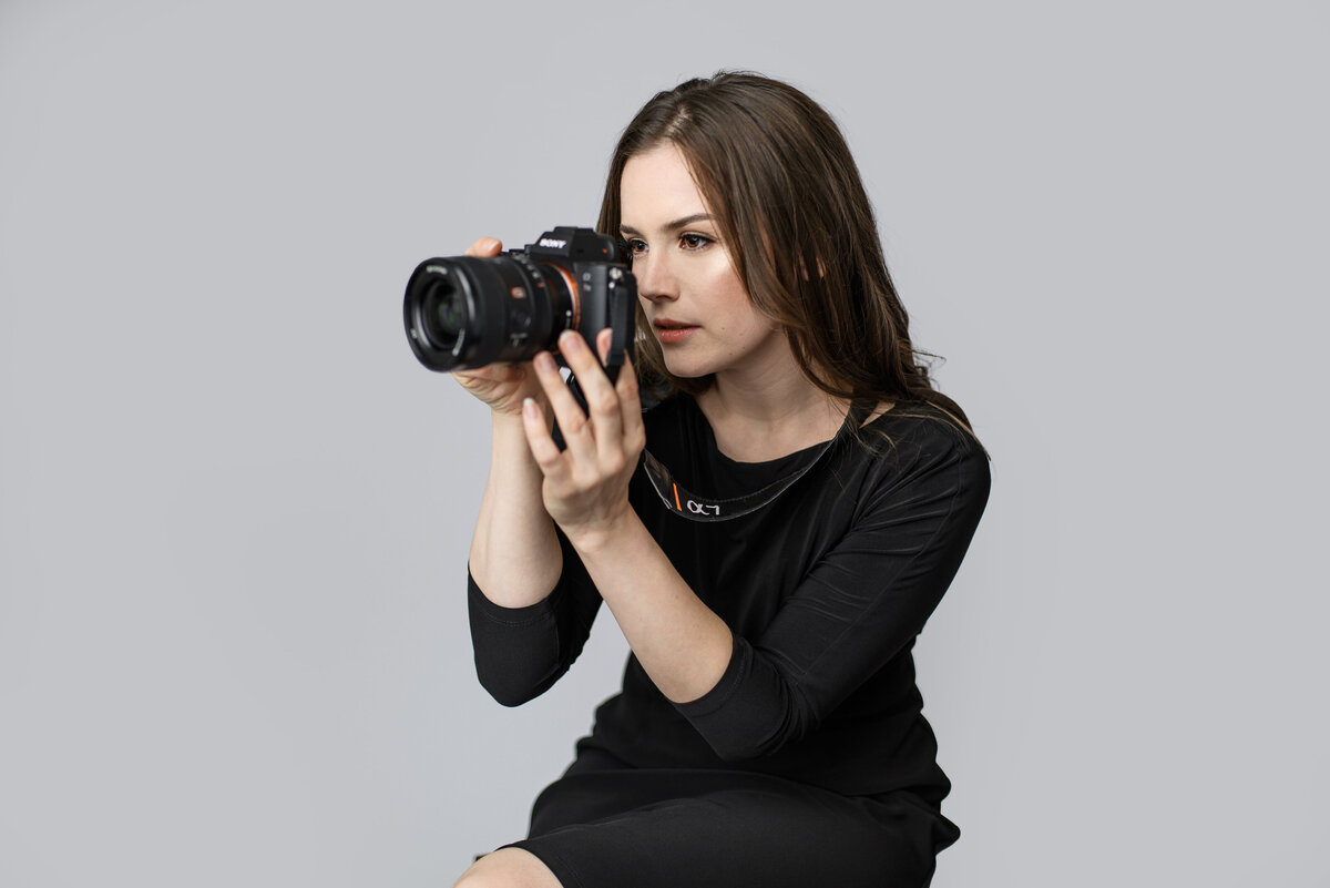Lana Stewart photographer holding camera and directing client while looking into viewfinder wearing black dress on gray background