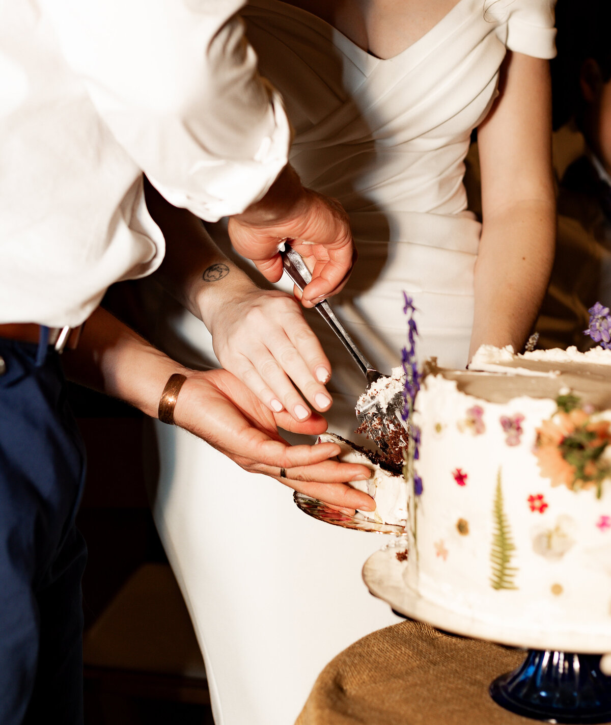 A couple cuts a wedding cake adorned with colorful edible flowers, captured perfectly by a New England Wedding Photographer. The bride, in her white dress, and groom gently hold the knife together as the cake rests elegantly on a brown tablecloth.