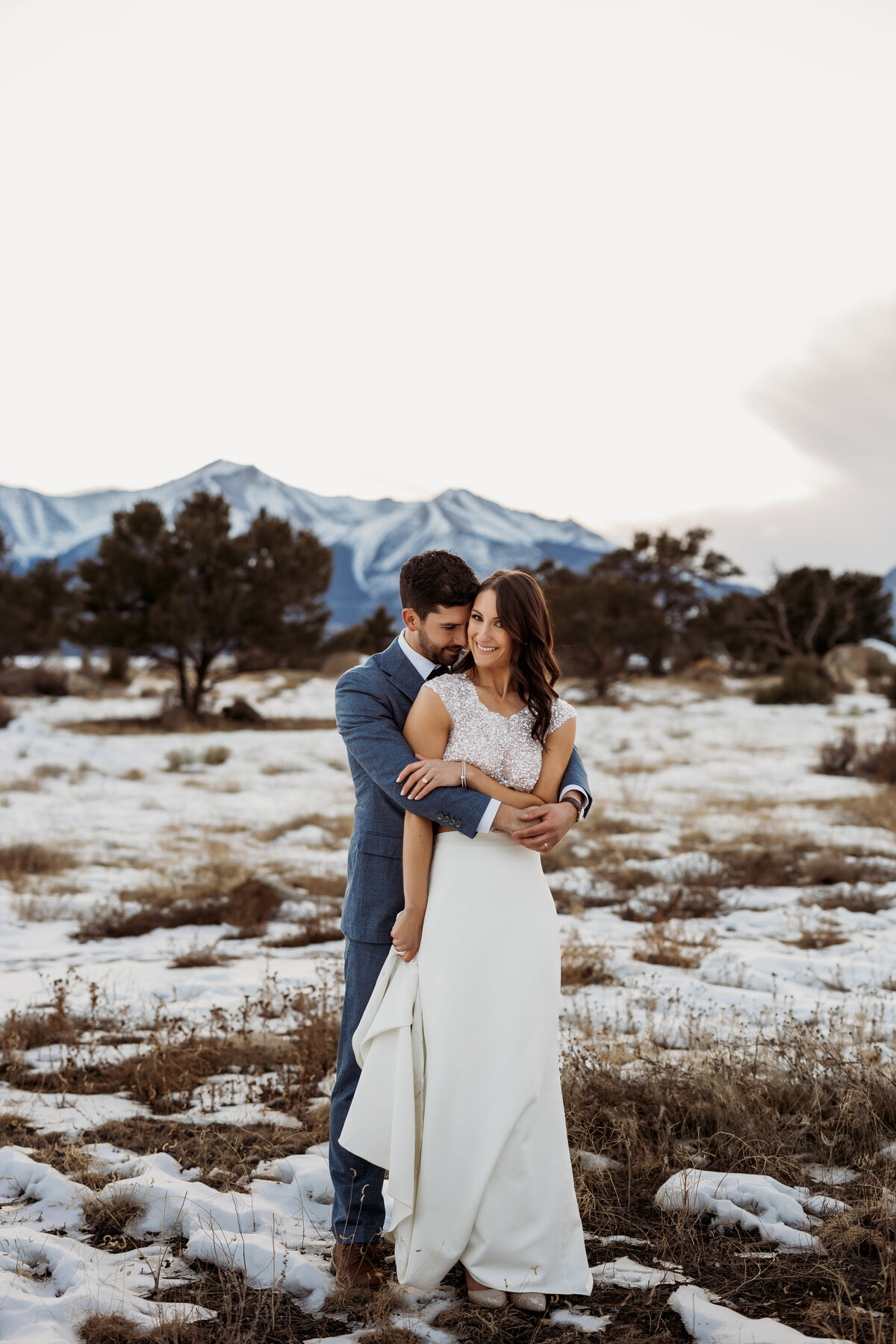 Husband embraces wife from behind while standing in a snowy field in front of snow capped mountains in Buena Vista, Colorado