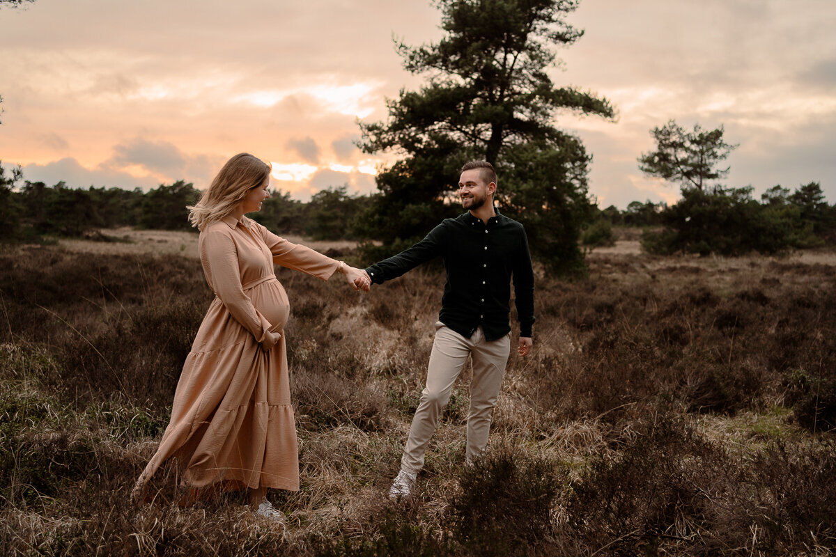 Man leidt zijn zwangere vrouw rustig over de heide tijdens een zwangerschapsshoot in de omgeving van Apeldoorn