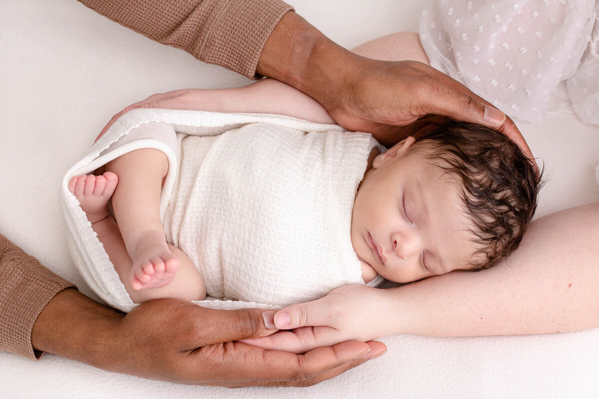 Baby swaddled in white sleeping on a white blanket and being  held by both parents with their arms holding each other and resting on the baby.