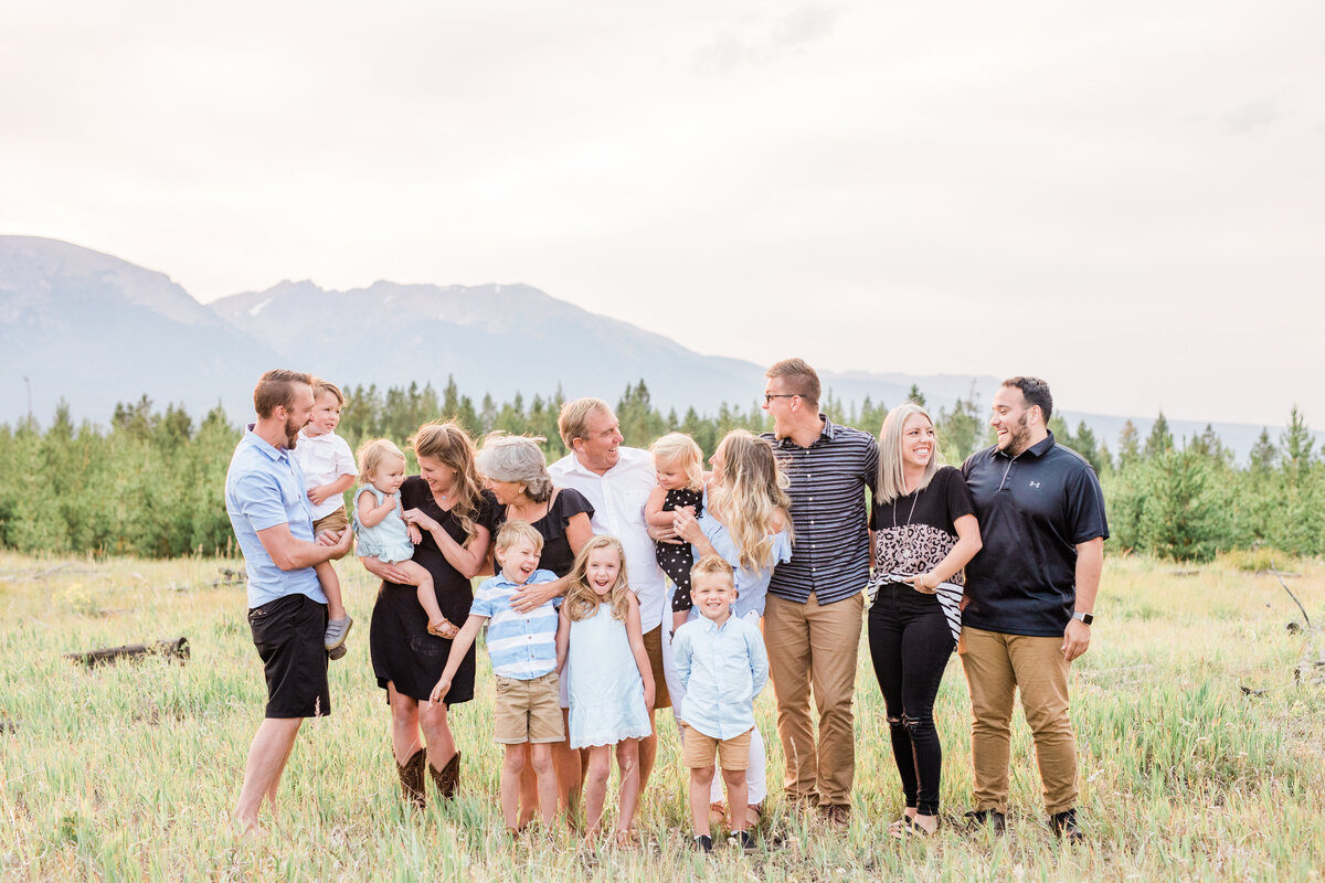 An extended family of 13 are all looking towards one another and laughing. They are in a grassy filed with pine trees and mountains behind then.