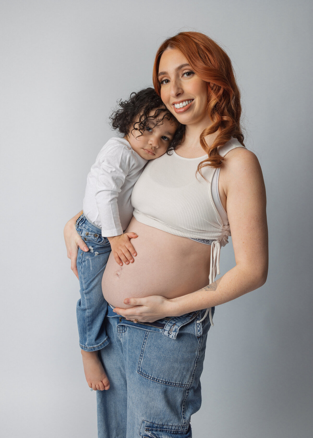 A tender family moment as a young child embraces a pregnant mother wearing a white crop top and jeans against a gray backdrop near Montclair, NJ.