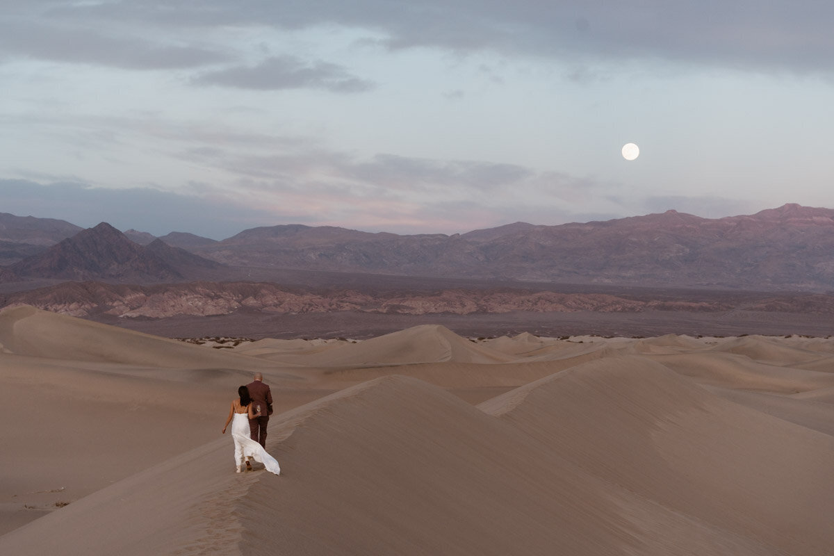 Bride and groom walking along the ridge of a sand dune with mountains and the moon in the backyard in Death Valley.