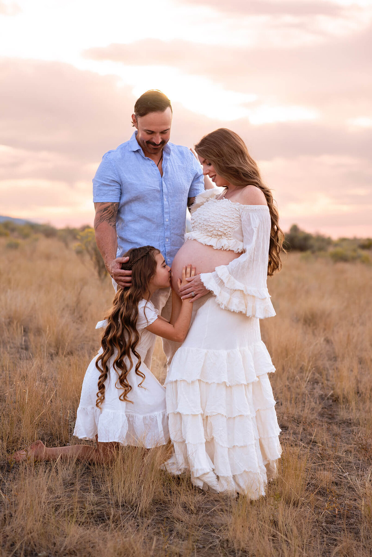 A family of 3 poses for a beautiful portrait. The mother and daughter both have long brown hair, wearing white. Husband is behind them and young daughter kisses moms belly.