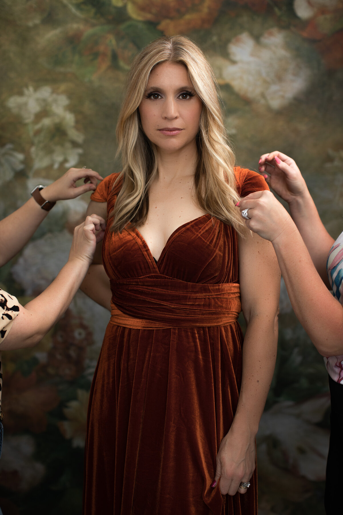 A blonde woman in a burnt orange dress is tended to and pampered as she poses for a portrait on a flower backdrop at Janel Lee Photography studios in Cincinnati Ohio