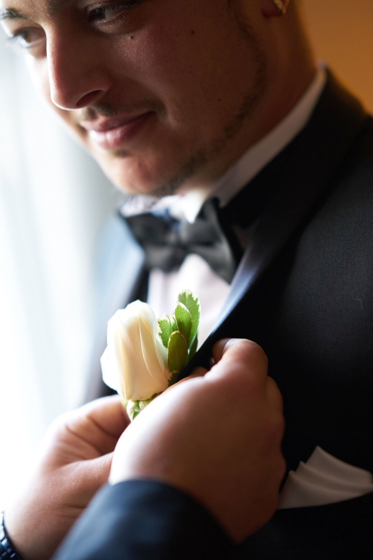 A close-up of someone pinning a boutonnière to the groom’s lapel, capturing a detailed moment of the finishing touches on his wedding attire. This image highlights the elegance of the groom’s outfit and the meticulous care taken to complete his look for the special day.