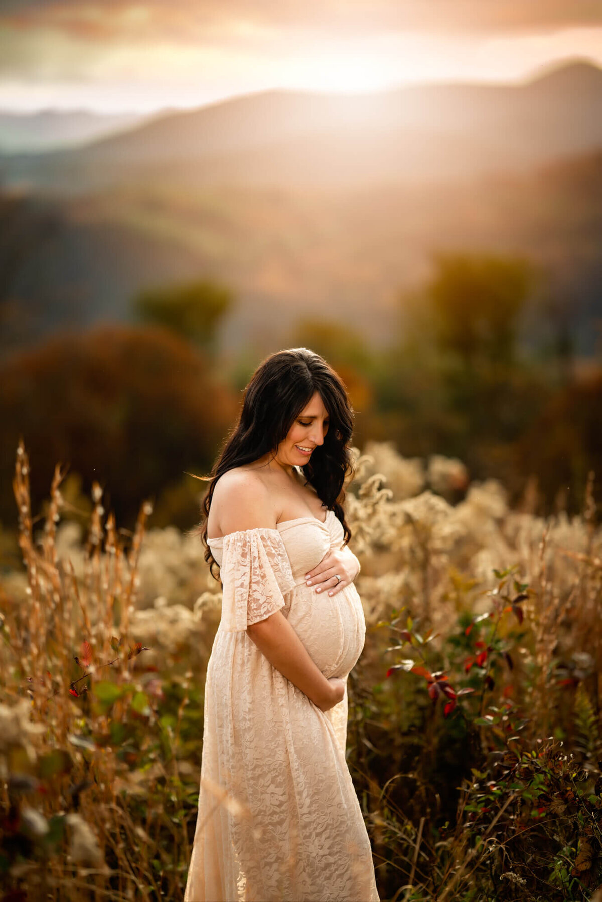 A dark haired mama to be in a long cream dress cradles her baby bump