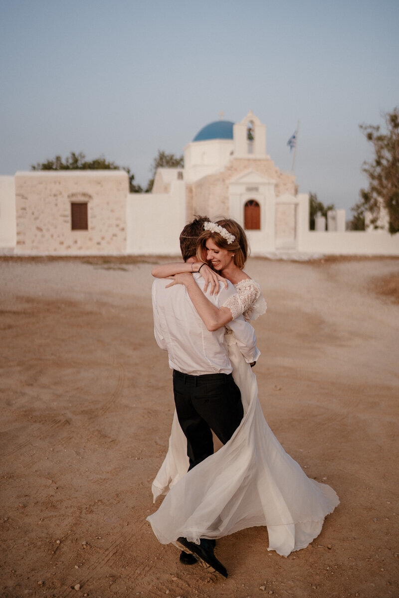 Couple de mariés devant l’église Agios Fokas à Paros, en Grèce, un moment symbolique et lumineux d’un mariage à l’étranger.
