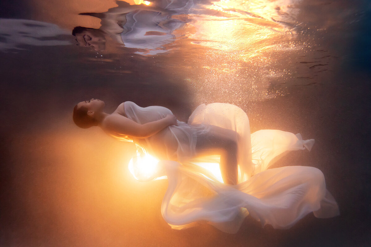 Dreamlike underwater photoshoot featuring an elegant woman enveloped in flowing white fabric, illuminated by a golden glow from beneath. Captured by a Dallas underwater pregnancy photographer, the scene evokes warmth, peace, and radiant beauty. The gentle motion of the fabric and ethereal light create a captivating, serene composition.
