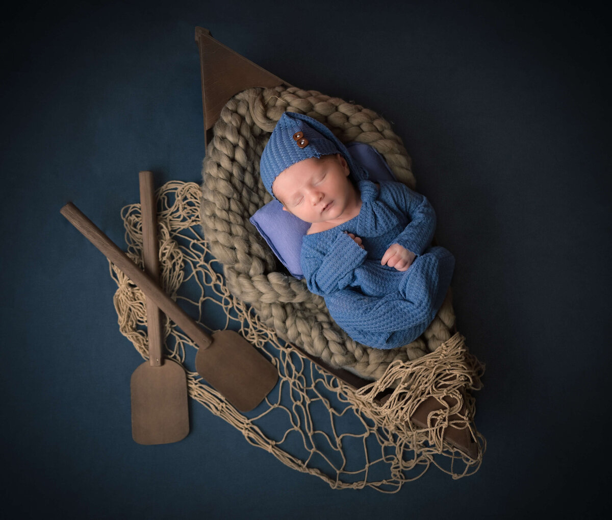 In-home newborn session of a baby posed sleeping in a row boat on a blue backdrop, fishing net, and oars.