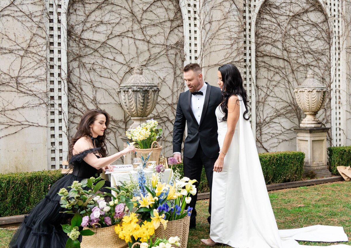 A live illustrator paints at a wedding while the bride and groom watch, surrounded by flowers in an elegant outdoor setting.