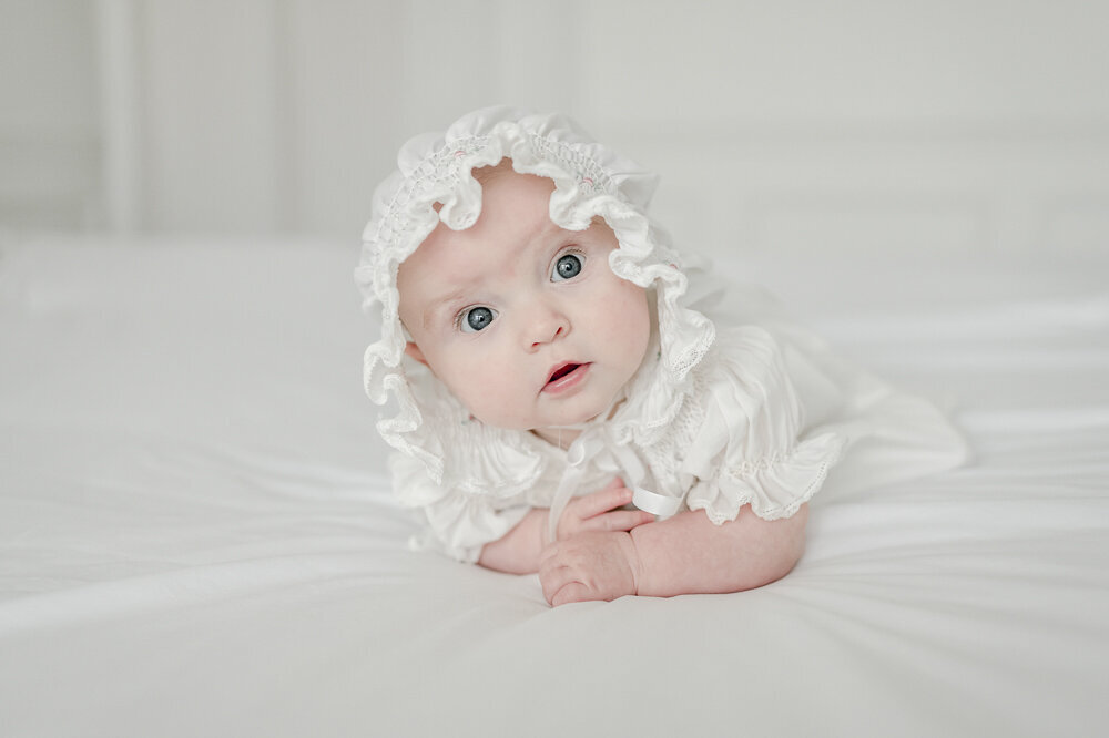 3 month old baby girl in a white ruffle bonnet lays on a white bed in in kristie lloyd's nashville photography studio