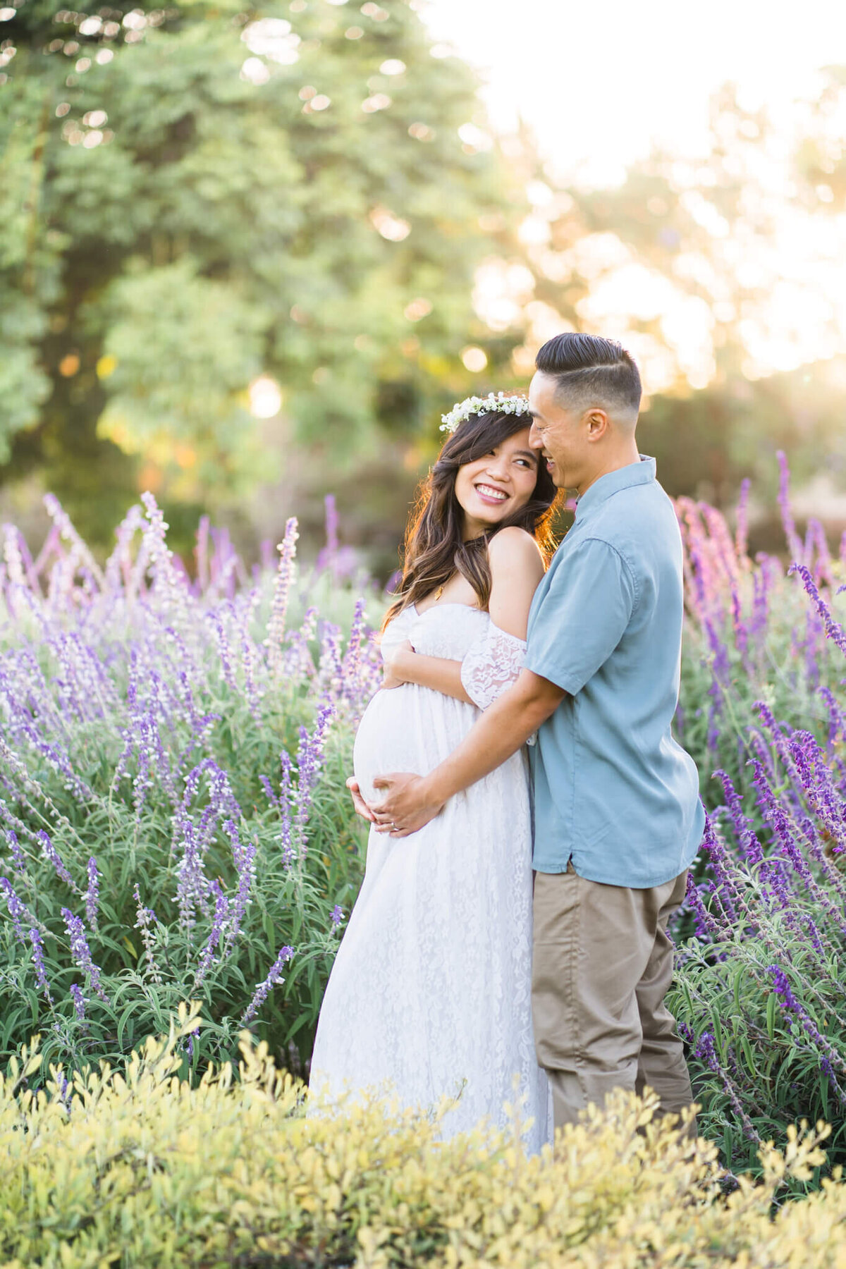 Pregnant couple hugging in a field of purple flowers with white flower wreath at golden hour