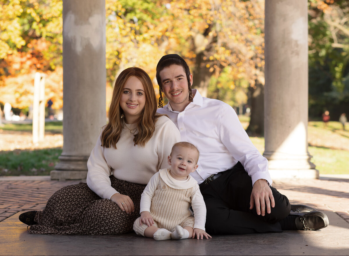 Mom, dad, and baby pose for a first birthday family portrait at a brooklyn, ny park. they are sitting under a stone portico and all are smiling at the camera. Captured by premier Brooklyn NY family photographer Chaya Bornstein Photography.
