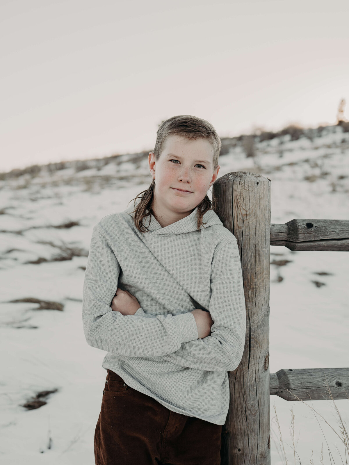 A young boy leans up against an old farm fence post with snow in the background as he confidently fold his arms in front of his body, while smiling at the camera, wearing a gray sweater.